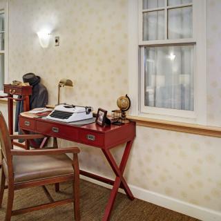 A vintage office setup with a typewriter on a red desk, accompanied by a wooden chair and a nearby bookshelf.