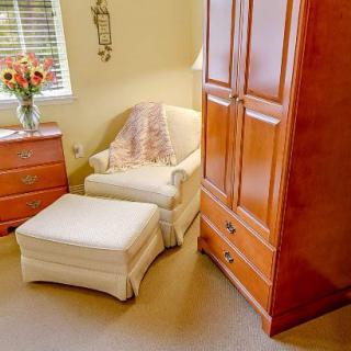 A cozy living room corner with an armchair and ottoman, next to a wooden cabinet and a vase of flowers by the window.