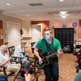A musician performs for residents at a nursing home, playing guitar and wearing a mask.
