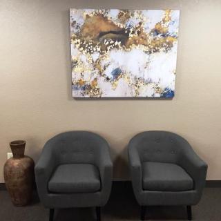A waiting area with two gray chairs, a decorative vase, and a painting on the wall.