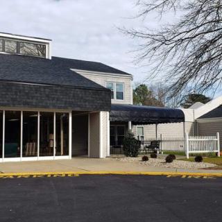 A commercial building with large windows and an awning, adjacent to a parking lot and a tree.