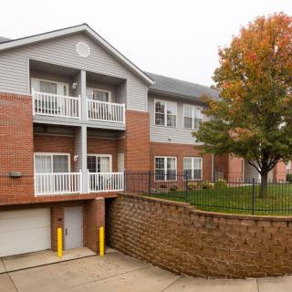 A two-story apartment building with balconies and a garage, surrounded by a brick retaining wall and a tree with autumn foliage.