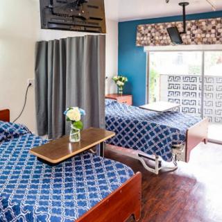 A hospital room with two beds featuring blue bedding and a table with flowers near a window.