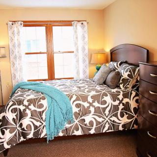 A cozy bedroom with a patterned bedspread and a teal blanket, featuring a window with curtains and wooden furniture.