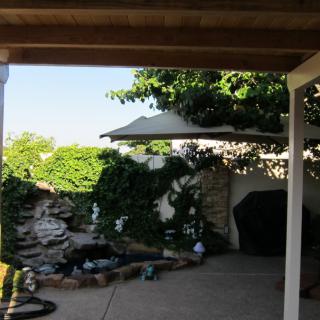 A tranquil garden patio featuring a small pond and waterfall, surrounded by lush ivy and shaded by a canopy.