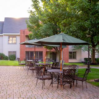 An outdoor patio area with tables and chairs under umbrellas, surrounded by greenery and a brick pathway.