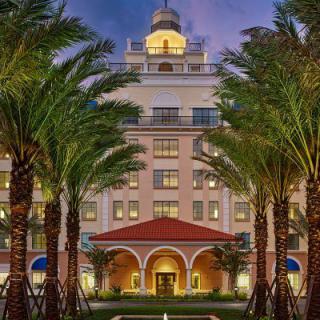A luxurious hotel building beautifully lit in the evening, framed by tall palm trees.