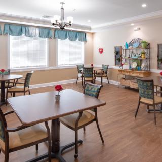 A cozy dining room with neatly arranged tables and chairs, featuring large windows with curtains and a chandelier.