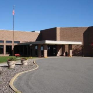 The image shows the entrance of a brick school building with a flagpole and parking area in front.