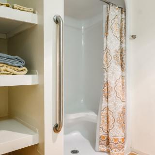 A bathroom with a shower, complete with a grab bar and patterned shower curtain. Towels are neatly stored on shelves beside the shower.