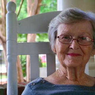 An elderly woman wearing glasses sits comfortably on a porch rocking chair, smiling warmly.