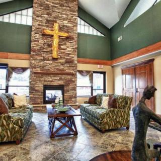 A spacious living room with a high ceiling features a stone fireplace adorned with a wooden cross, surrounded by cozy sofas and large windows.