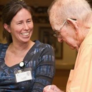 A woman smiling while engaging in a conversation with an elderly person indoors.