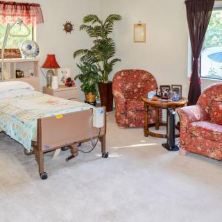 A cozy bedroom with a hospital bed and comfortable seating area, featuring two armchairs and a small table near a large window.