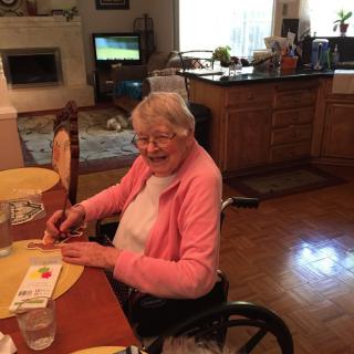 An elderly woman in a wheelchair sitting at a dining table in a cozy home setting.