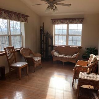 A cozy living room with wooden floors illuminated by sunlight through large windows, featuring various chairs and a sofa.