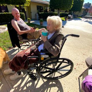 An elderly woman enjoying a sunny day outside, conversing with a man while seated in her wheelchair.