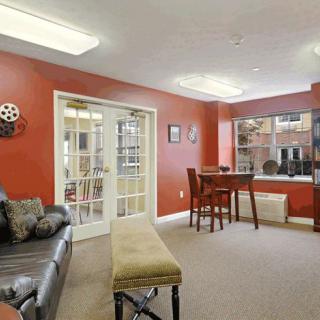 A cozy living room with red walls, featuring a comfortable sofa, a small table with chairs by the window, and a decorative wall piece.