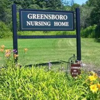 A sign for Greensboro Nursing Home surrounded by greenery and flowers.
