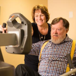 An elderly man receives assistance while using exercise equipment.