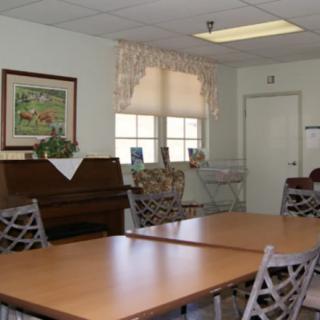 A cozy room with a piano, tables, and chairs, decorated with artwork and natural light from the window.
