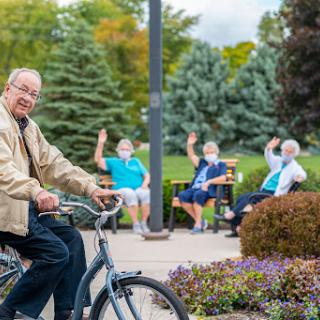 A cheerful man enjoying a bike ride in the park, greeted by friendly seniors sitting nearby.