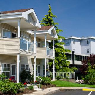 A sunny view of two residential apartment buildings with balconies, surrounded by trees and landscaping.