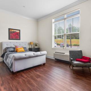 A modern bedroom with wooden flooring, featuring a bed, armchair, and large window allowing natural light.