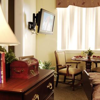 A cozy room featuring a television mounted on the wall, a wooden table with chairs, and a lamp on a dresser.