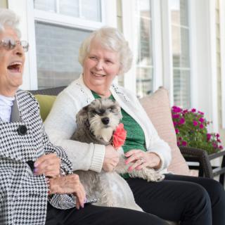 Two elderly women sitting outside, smiling and enjoying time with a small dog.