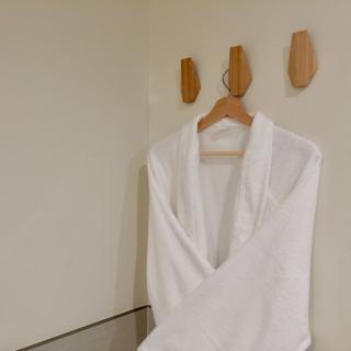 A white bathrobe hangs on a wooden hanger against a minimalist wall with wooden hooks.