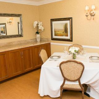 A warmly decorated dining room with a table set for two, featuring elegant chairs, a painting on the wall, and a large mirror above a sideboard.