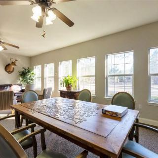 A well-lit dining room with a wooden table, surrounded by chairs, and illuminated by natural light through large windows.