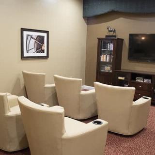 A cozy waiting room with beige chairs facing a television, accompanied by artwork on the wall and a bookshelf in the corner.