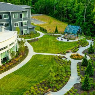 Aerial view of a landscaped garden with pathways, a building, and a small greenhouse surrounded by trees.