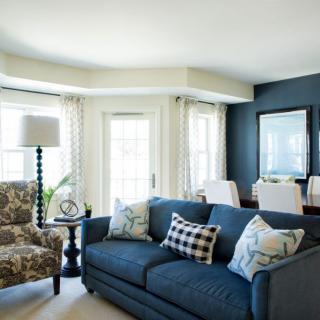 A cozy living room with a blue sofa, patterned armchair, and a bright dining area in the background.