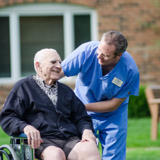 A caregiver spending time with an elderly person in a wheelchair outdoors.