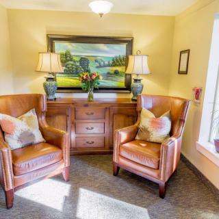 A cozy corner with two armchairs and a sideboard, adorned with lamps and a scenic painting.