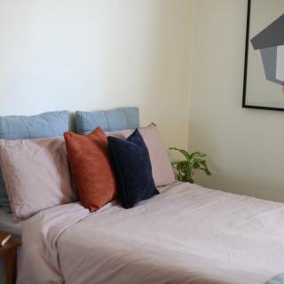 A neatly arranged bedroom with a bed adorned with colorful pillows, accompanied by artwork and a plant.
