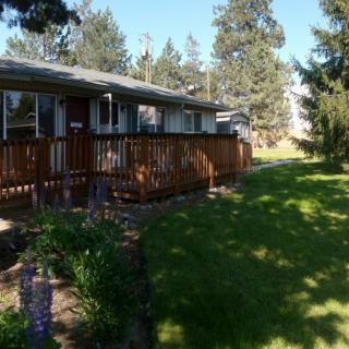 A single-story house with a wooden porch, surrounded by a lush green lawn and trees.