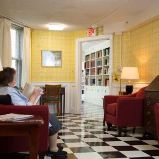 A cozy living room with yellow walls, red chairs, and a checkerboard floor. A person is seated, reading a book.