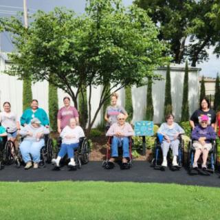 A group of people sitting in wheelchairs outdoors in a garden setting.
