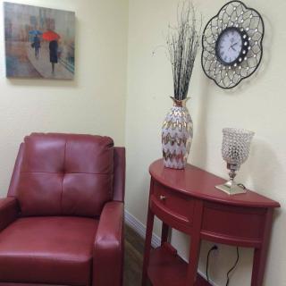 A cozy corner featuring a red armchair and matching side table, accented with a vase, wall art, and a decorative clock.