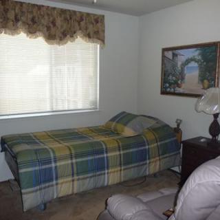 A small bedroom with a single bed covered in a plaid blanket, a window with patterned curtains, and a framed painting on the wall.