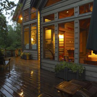 A cozy wooden house with a warmly lit interior, viewed from a rain-soaked porch in the evening.