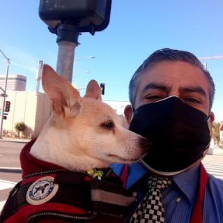 A man wearing a mask poses for a selfie with his dog on the street.