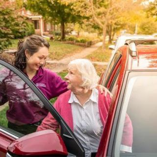 A young woman assists an elderly woman getting out of a car in an outdoor setting.