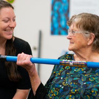 An elderly woman lifting a blue bar while being encouraged by a younger woman.