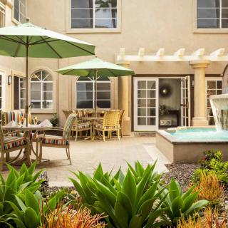 A cozy outdoor patio area with green umbrellas, tables, and lush plants surrounding a fountain.