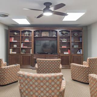 A cozy living room with armchairs facing a large television, surrounded by built-in bookshelves and lit by skylights.